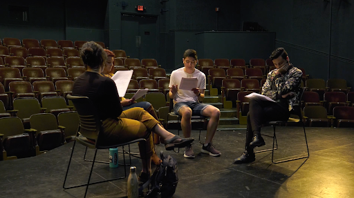 Students sitting on a theater stage reading from scripts