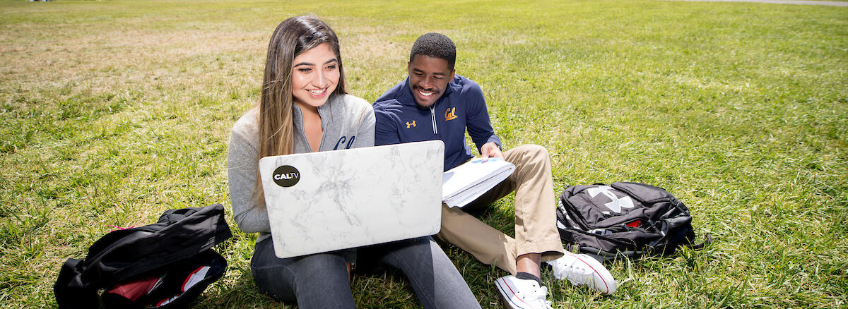 Two students sit on a grassy lawn at UC Berkeley, smiling and looking at a laptop.