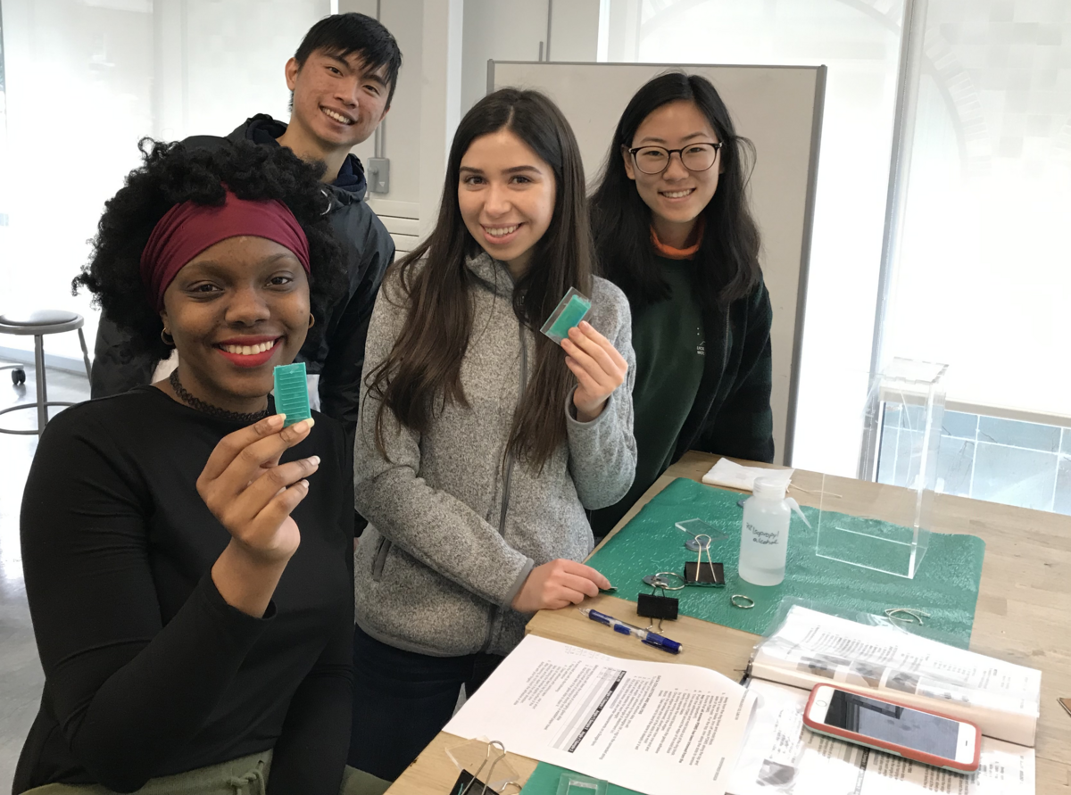 Four students smile at the camera while holding a green material. The same green material, a textbook, a phone, papers, pens, clips, and isopropyl alcohol are spread out over a worktable to the right.